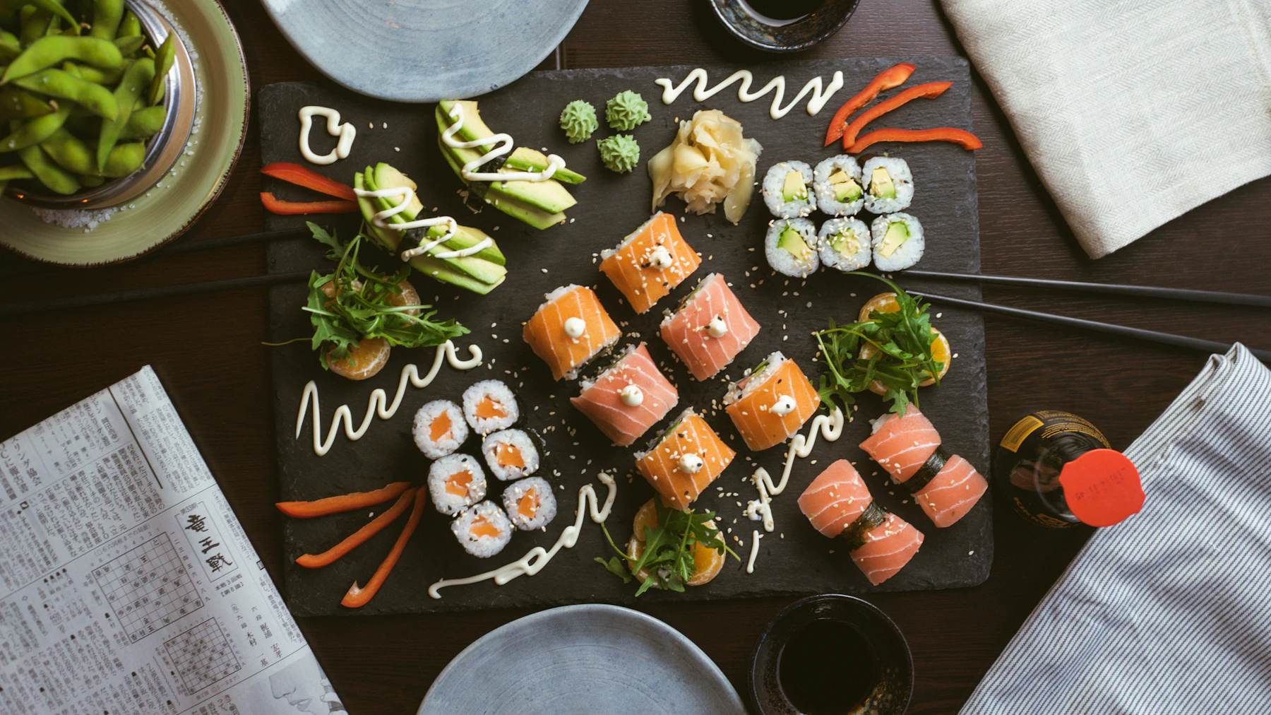 Assorted sushi rolls on a slate platter with edamame, chopsticks, and table settings